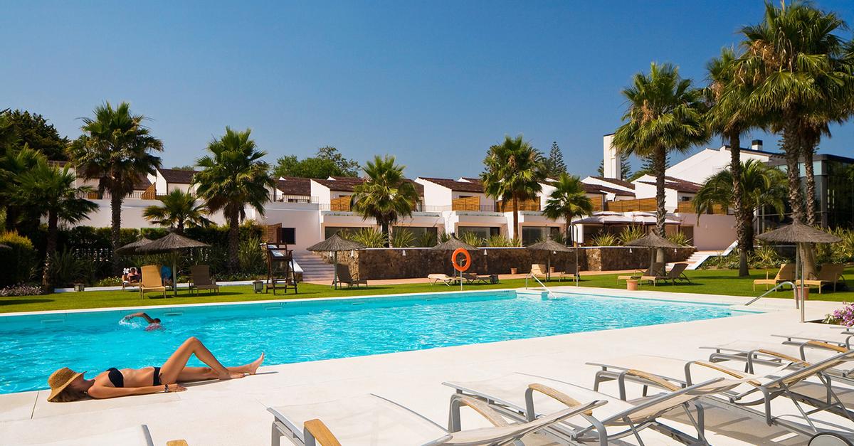 Una pintoresca escena de piscina al aire libre en Sotogrande cuenta con tumbonas y una persona descansando entre palmeras, bajo un cielo despejado y en medio de majestuosos edificios blancos: una escapada de fin de semana perfecta con una elegante cena bajo las estrellas, complementada con una botella de cava. - ForTwo - Planes para parejas