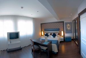 Habitación en el Hotel Gran Versalles Madrid con cama de matrimonio, escritorio, TV y grandes ventanales con cortinas transparentes. - ForTwo - Planes para parejas