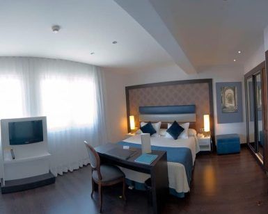 Habitación en el Hotel Gran Versalles Madrid con cama de matrimonio, escritorio, TV y grandes ventanales con cortinas transparentes. - ForTwo - Planes para parejas