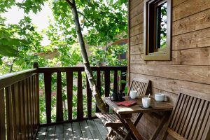 La Habitación Casa del árbol Maitenea del Hotel Jakue (Navarra) incluye un patio de madera con mesa, sillas, tazas y un cuaderno rodeado de frondosos árboles verdes que crean un ambiente tranquilo. - ForTwo - Planes para parejas