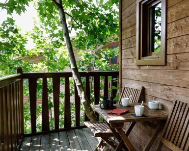 La Habitación Casa del árbol Maitenea del Hotel Jakue (Navarra) incluye un patio de madera con mesa, sillas, tazas y un cuaderno rodeado de frondosos árboles verdes que crean un ambiente tranquilo. - ForTwo - Planes para parejas