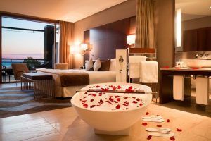 Habitación de lujo en el Hotel Miramar Barcelona GL, con bañera adornada con pétalos de rosa, cama de felpa y una impresionante vista al mar al atardecer. - ForTwo - Planes para parejas