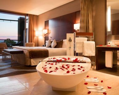 Habitación de lujo en el Hotel Miramar Barcelona GL, con bañera adornada con pétalos de rosa, cama de felpa y una impresionante vista al mar al atardecer. - ForTwo - Planes para parejas