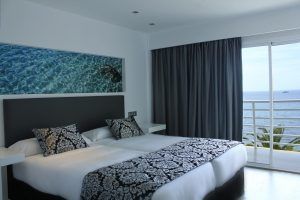 Disfrute de la comodidad del Hotel Náutico Ebeso en Ibiza con una habitación que ofrece dos camas, ropa de cama estampada elegante, un balcón con vista al mar y una impresionante obra de arte de agua azul sobre el cabecero. - ForTwo - Planes para parejas