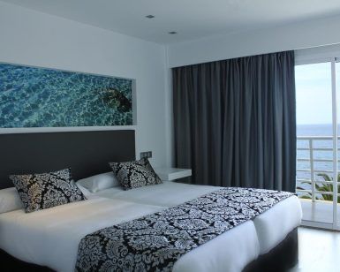 Disfrute de la comodidad del Hotel Náutico Ebeso en Ibiza con una habitación que ofrece dos camas, ropa de cama estampada elegante, un balcón con vista al mar y una impresionante obra de arte de agua azul sobre el cabecero. - ForTwo - Planes para parejas