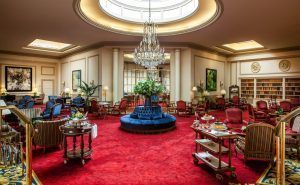 El lujoso lobby del Hotel Wellington Madrid exhibe una alfombra roja, elegantes asientos y lámparas de araña bajo un gran tragaluz adornado con plantas vibrantes. - ForTwo - Planes para parejas