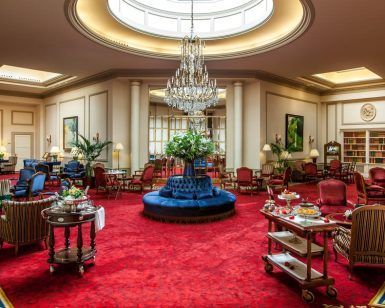 El lujoso lobby del Hotel Wellington Madrid exhibe una alfombra roja, elegantes asientos y lámparas de araña bajo un gran tragaluz adornado con plantas vibrantes. - ForTwo - Planes para parejas