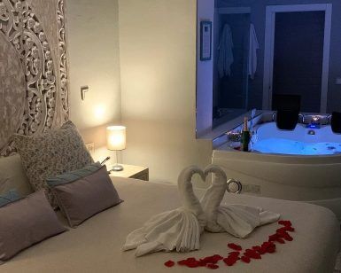 Experimente la Escapada Romántica en Suite con Jacuzzi en el Hotel Adealba, Mérida, con toallas de cisne adornando la cama y un rastro de pétalos de rosa que conduce a su bañera de hidromasaje privada iluminada en la esquina. - ForTwo - Planes para parejas