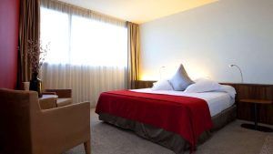 Disfrute de una Noche Deluxe con desayuno y cava en SB Diagonal Zero. Relájese en nuestra moderna habitación, con una cama grande con manta roja, sillón, mesitas de noche y elegantes ventanas con cortinas beige, creando el ambiente perfecto para relajarse. - ForTwo - Planes para parejas