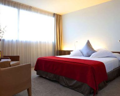 Disfrute de una Noche Deluxe con desayuno y cava en SB Diagonal Zero. Relájese en nuestra moderna habitación, con una cama grande con manta roja, sillón, mesitas de noche y elegantes ventanas con cortinas beige, creando el ambiente perfecto para relajarse. - ForTwo - Planes para parejas
