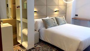 Experimente una moderna habitación de hotel con estanterías abiertas, una elegante cama, suelo estampado, apliques de pared y vistas increíbles del encanto de Barcelona con Escapada en hotel 4 estrellas. - ForTwo - Planes para parejas