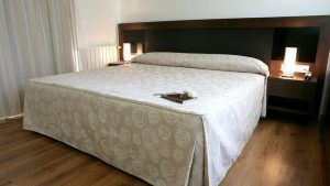 Una cama cuidadosamente hecha con ropa de cama con diseño en espiral, dos mesas de noche con lámparas encendidas y un piso de madera pulida crean un ambiente sereno que recuerda a la experiencia de Escapada en el Hotel Sinagoga con desayuno, bombones y spa (Hervás). - ForTwo - Planes para parejas