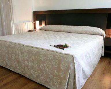 Una cama cuidadosamente hecha con ropa de cama con diseño en espiral, dos mesas de noche con lámparas encendidas y un piso de madera pulida crean un ambiente sereno que recuerda a la experiencia de Escapada en el Hotel Sinagoga con desayuno, bombones y spa (Hervás). - ForTwo - Planes para parejas