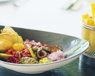 En el romántico Restaurante Ibiza Hoyo 19 se sirve un plato gourmet con tartar, patatas fritas, alcaparras y cebollas elegantemente presentadas con patatas fritas en una cesta de alambre. - ForTwo - Planes para parejas
