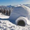 Un iglú nevado se yergue majestuoso ante un telón de fondo de montañas y bosques bajo un cielo azul despejado, evocando la serenidad de los paisajes cercanos a Navacerrada. Vívelo con "Construye un iglú en Navacerrada (Madrid)". - ForTwo - Planes para parejas