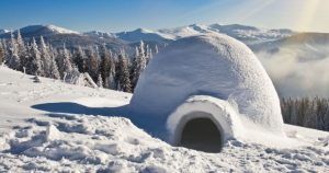 Un iglú nevado se yergue majestuoso ante un telón de fondo de montañas y bosques bajo un cielo azul despejado, evocando la serenidad de los paisajes cercanos a Navacerrada. Vívelo con "Construye un iglú en Navacerrada (Madrid)". - ForTwo - Planes para parejas