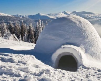 Un iglú nevado se yergue majestuoso ante un telón de fondo de montañas y bosques bajo un cielo azul despejado, evocando la serenidad de los paisajes cercanos a Navacerrada. Vívelo con "Construye un iglú en Navacerrada (Madrid)". - ForTwo - Planes para parejas