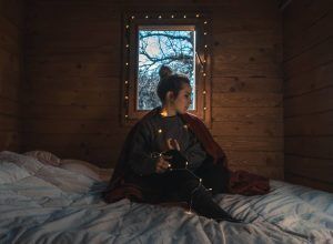 Dentro de una Cabaña Águila Imperial en el Parque Nacional de Cabañeros, una mujer está sentada en una cama con luces de cadena. Envuelta en un suéter, contempla el impresionante paisaje del parque. - ForTwo - Planes para parejas