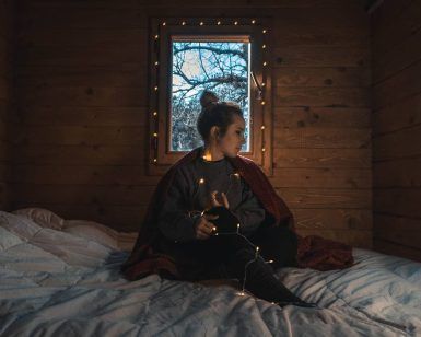 Dentro de una Cabaña Águila Imperial en el Parque Nacional de Cabañeros, una mujer está sentada en una cama con luces de cadena. Envuelta en un suéter, contempla el impresionante paisaje del parque. - ForTwo - Planes para parejas