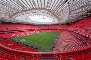 El majestuoso Estadio San Mamés de Bilbao se encuentra vacío con sus asientos rojos contrastando con el exuberante campo verde bajo un gran techo ovalado retráctil, esperando ansiosamente la vitalidad del próximo Tour por el estadio San Mamés (Bilbao). - ForTwo - Planes para parejas