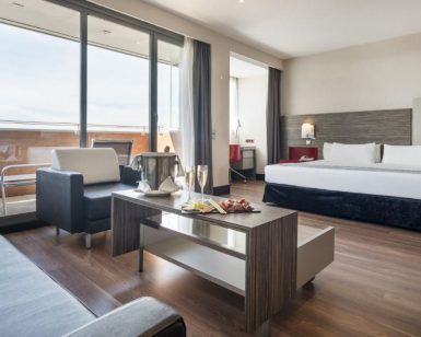 Descubre la elegancia de la Junior Suite: el secreto de Ilunion Hotels (Barcelona), con un diseño moderno que incluye una amplia cama, acogedora zona de estar, balcón y una mesa preparada con snacks y champán frío. - ForTwo - Planes para parejas