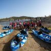 Un grupo con chalecos salvavidas y remos preparados se encuentra en la tranquila orilla del lago Sevilla, rodeado de kayaks azules. Este momento queda plasmado en la aventurera película "Kayak con reportaje fotográfico" (Sevilla), que destaca la emoción y la camaradería del kayak. - ForTwo - Planes para parejas