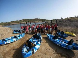 Un grupo con chalecos salvavidas y remos preparados se encuentra en la tranquila orilla del lago Sevilla, rodeado de kayaks azules. Este momento queda plasmado en la aventurera película "Kayak con reportaje fotográfico" (Sevilla), que destaca la emoción y la camaradería del kayak. - ForTwo - Planes para parejas