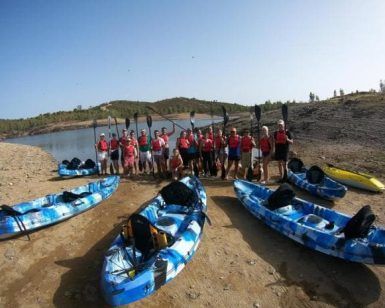 Un grupo con chalecos salvavidas y remos preparados se encuentra en la tranquila orilla del lago Sevilla, rodeado de kayaks azules. Este momento queda plasmado en la aventurera película "Kayak con reportaje fotográfico" (Sevilla), que destaca la emoción y la camaradería del kayak. - ForTwo - Planes para parejas