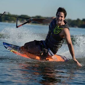 Un hombre sonriente practica wakeboard con destreza en un lago, creando un deslumbrante chorro de agua al girar. Enmarcado por frondosos árboles al fondo, su estilo refleja la vibrante energía de "Una forma diferente de surfear, descubre el Kneeboard en Barcelona". - ForTwo - Planes para parejas