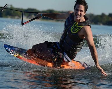 Un hombre sonriente practica wakeboard con destreza en un lago, creando un deslumbrante chorro de agua al girar. Enmarcado por frondosos árboles al fondo, su estilo refleja la vibrante energía de "Una forma diferente de surfear, descubre el Kneeboard en Barcelona". - ForTwo - Planes para parejas