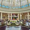 El elegante vestíbulo del Restaurante La Rotonda en el Hotel Westin Palace Madrid exhibe un techo de cristal abovedado, una lámpara de araña deslumbrante, sillas coloridas y un impresionante arreglo floral central. - ForTwo - Planes para parejas