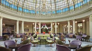 El elegante vestíbulo del Restaurante La Rotonda en el Hotel Westin Palace Madrid exhibe un techo de cristal abovedado, una lámpara de araña deslumbrante, sillas coloridas y un impresionante arreglo floral central. - ForTwo - Planes para parejas