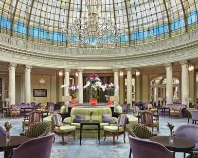 El elegante vestíbulo del Restaurante La Rotonda en el Hotel Westin Palace Madrid exhibe un techo de cristal abovedado, una lámpara de araña deslumbrante, sillas coloridas y un impresionante arreglo floral central. - ForTwo - Planes para parejas