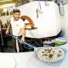 En la rústica cocina del Restaurante Leccabaffi, el chef coloca una pizza cruda en el horno de ladrillo, sonriendo bajo su gorra y delantal durante "Martes de música en vivo y pizza". - ForTwo - Planes para parejas
