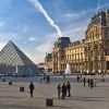 La pirámide del Louvre y el museo histórico de París se alzan majestuosos bajo un cielo azul mientras los visitantes disfrutan de la Visita guiada al Louvre (París) alrededor de la bulliciosa plaza. - ForTwo - Planes para parejas