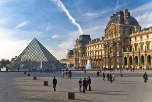 La pirámide del Louvre y el museo histórico de París se alzan majestuosos bajo un cielo azul mientras los visitantes disfrutan de la Visita guiada al Louvre (París) alrededor de la bulliciosa plaza. - ForTwo - Planes para parejas