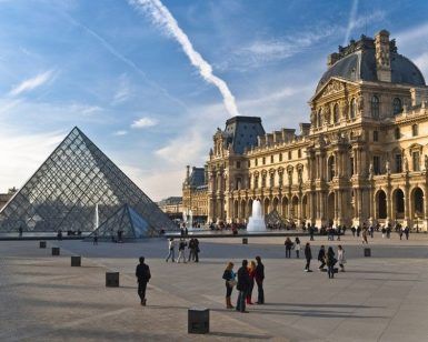 La pirámide del Louvre y el museo histórico de París se alzan majestuosos bajo un cielo azul mientras los visitantes disfrutan de la Visita guiada al Louvre (París) alrededor de la bulliciosa plaza. - ForTwo - Planes para parejas