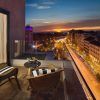 El acogedor balcón del Hotel H10 Puerta de Alcalá Madrid cuenta con sillas de mimbre y ofrece una vista impresionante del vibrante paisaje urbano y del cielo del atardecer sobre una bulliciosa calle. - ForTwo - Planes para parejas