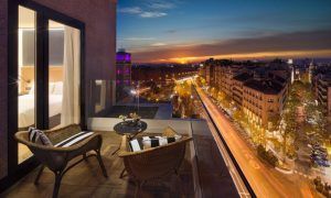 El acogedor balcón del Hotel H10 Puerta de Alcalá Madrid cuenta con sillas de mimbre y ofrece una vista impresionante del vibrante paisaje urbano y del cielo del atardecer sobre una bulliciosa calle. - ForTwo - Planes para parejas