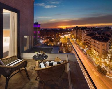 El acogedor balcón del Hotel H10 Puerta de Alcalá Madrid cuenta con sillas de mimbre y ofrece una vista impresionante del vibrante paisaje urbano y del cielo del atardecer sobre una bulliciosa calle. - ForTwo - Planes para parejas