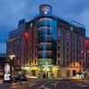El Hotel Santo Domingo Madrid, un moderno establecimiento con ventanas con marcos rojos, se ilumina bellamente al anochecer en una esquina de Santo Domingo, rodeado de automóviles y motocicletas. - ForTwo - Planes para parejas