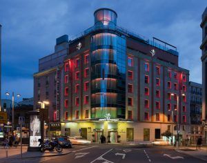 El Hotel Santo Domingo Madrid, un moderno establecimiento con ventanas con marcos rojos, se ilumina bellamente al anochecer en una esquina de Santo Domingo, rodeado de automóviles y motocicletas. - ForTwo - Planes para parejas