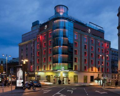 El Hotel Santo Domingo Madrid, un moderno establecimiento con ventanas con marcos rojos, se ilumina bellamente al anochecer en una esquina de Santo Domingo, rodeado de automóviles y motocicletas. - ForTwo - Planes para parejas