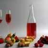 Un líquido rojo en dos vasos, una botella, frutas variadas y postres sobre una superficie blanca capturan la esencia de un picnic en Barcelona: una encarnación de colores vibrantes y verdadero deleite de "Picnic en Barcelona por sorpresa - Menú Dulce". - ForTwo - Planes para parejas