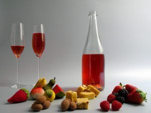 Un líquido rojo en dos vasos, una botella, frutas variadas y postres sobre una superficie blanca capturan la esencia de un picnic en Barcelona: una encarnación de colores vibrantes y verdadero deleite de "Picnic en Barcelona por sorpresa - Menú Dulce". - ForTwo - Planes para parejas