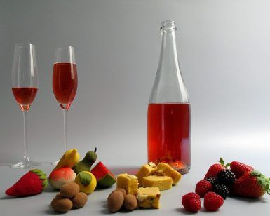 Un líquido rojo en dos vasos, una botella, frutas variadas y postres sobre una superficie blanca capturan la esencia de un picnic en Barcelona: una encarnación de colores vibrantes y verdadero deleite de "Picnic en Barcelona por sorpresa - Menú Dulce". - ForTwo - Planes para parejas