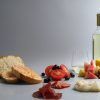 Disfrute de un delicioso Picnic en Barcelona - Menú Mar Salada con pan, tomates en rodajas, aceitunas, embutidos, fruta fresca y una botella de vino blanco sobre un encantador fondo gris. - ForTwo - Planes para parejas