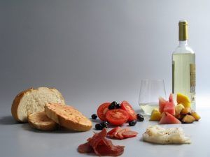 Disfrute de un delicioso Picnic en Barcelona - Menú Mar Salada con pan, tomates en rodajas, aceitunas, embutidos, fruta fresca y una botella de vino blanco sobre un encantador fondo gris. - ForTwo - Planes para parejas