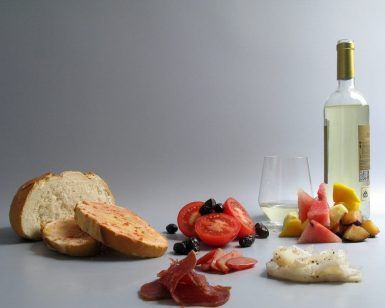 Disfrute de un delicioso Picnic en Barcelona - Menú Mar Salada con pan, tomates en rodajas, aceitunas, embutidos, fruta fresca y una botella de vino blanco sobre un encantador fondo gris. - ForTwo - Planes para parejas