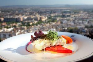 El plato de pescado gourmet, las verduras vibrantes y el paisaje urbano de fondo capturan perfectamente la esencia de Granada en el Hotel Alhambra Palace en el entorno "Almuerzo romántico a la carta". - ForTwo - Planes para parejas